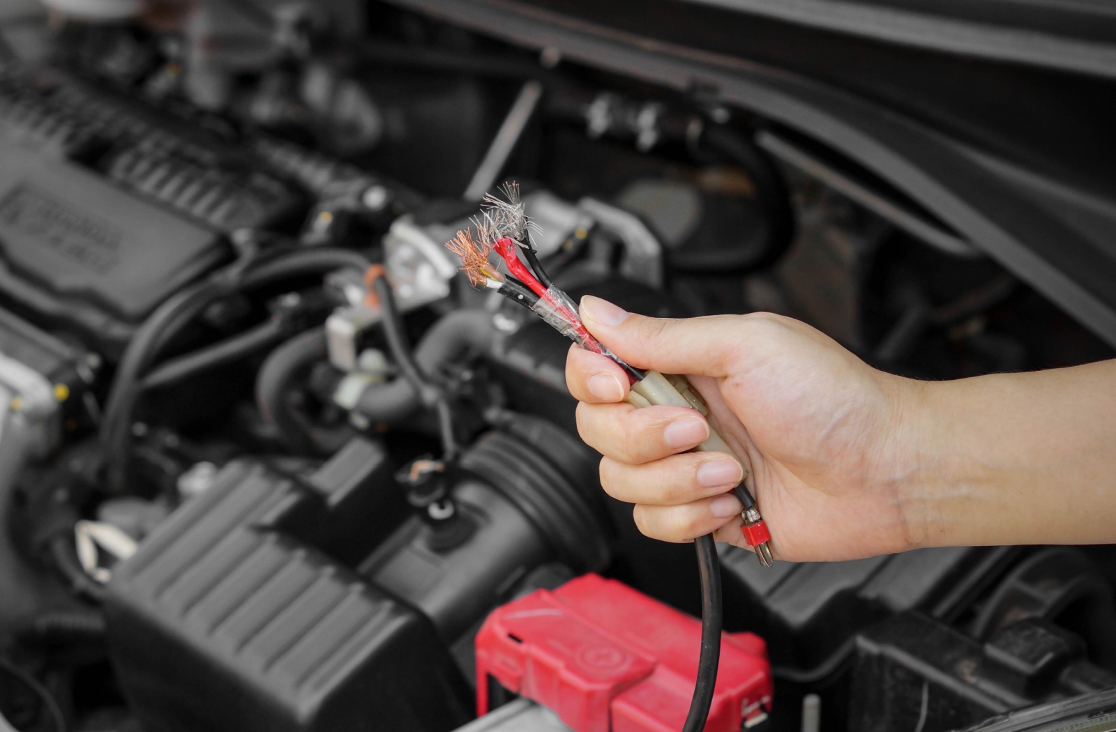 Hand holding damaged car wires in front of an open car engine, highlighting potential electrical issues.