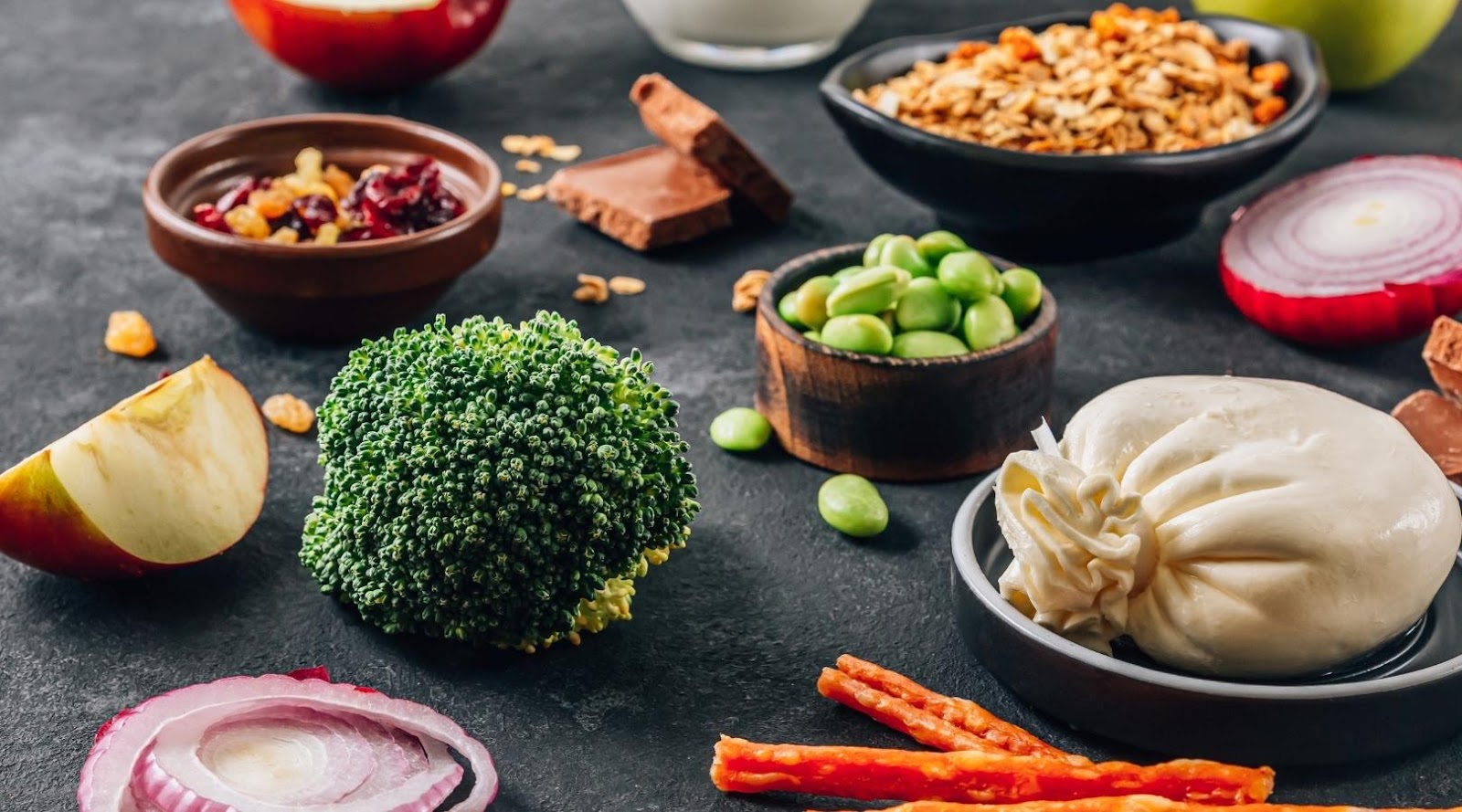 Fresh ingredients including broccoli, apple slices, red onion, edamame, burrata cheese, granola, and dried fruit on a dark surface.