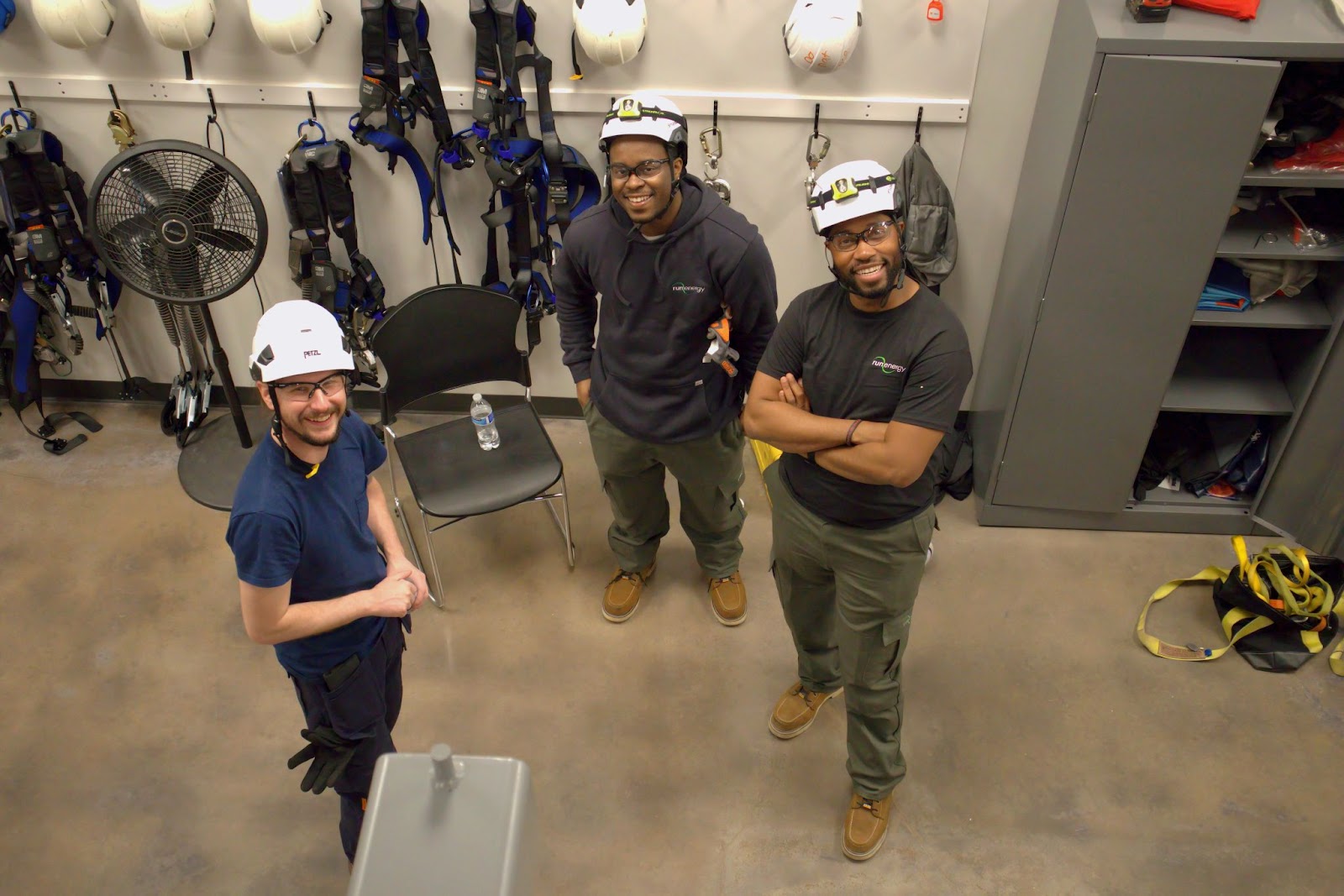 Three individuals wearing safety helmets and work attire smiling up at the camera in a training facility. Behind them are safety harnesses, helmets, and gear neatly organized on hooks and shelves, reflecting a professional and well-equipped environment.