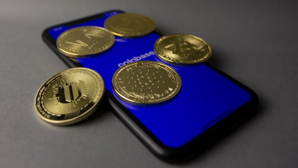 Mobile phone with a blue screen accessing Coinbase, surrounded by five cryptocurrencies: Polkadot, Solana, Bitcoin, Ethereum, and Cardano, symbolizing digital asset trading and portfolio management.