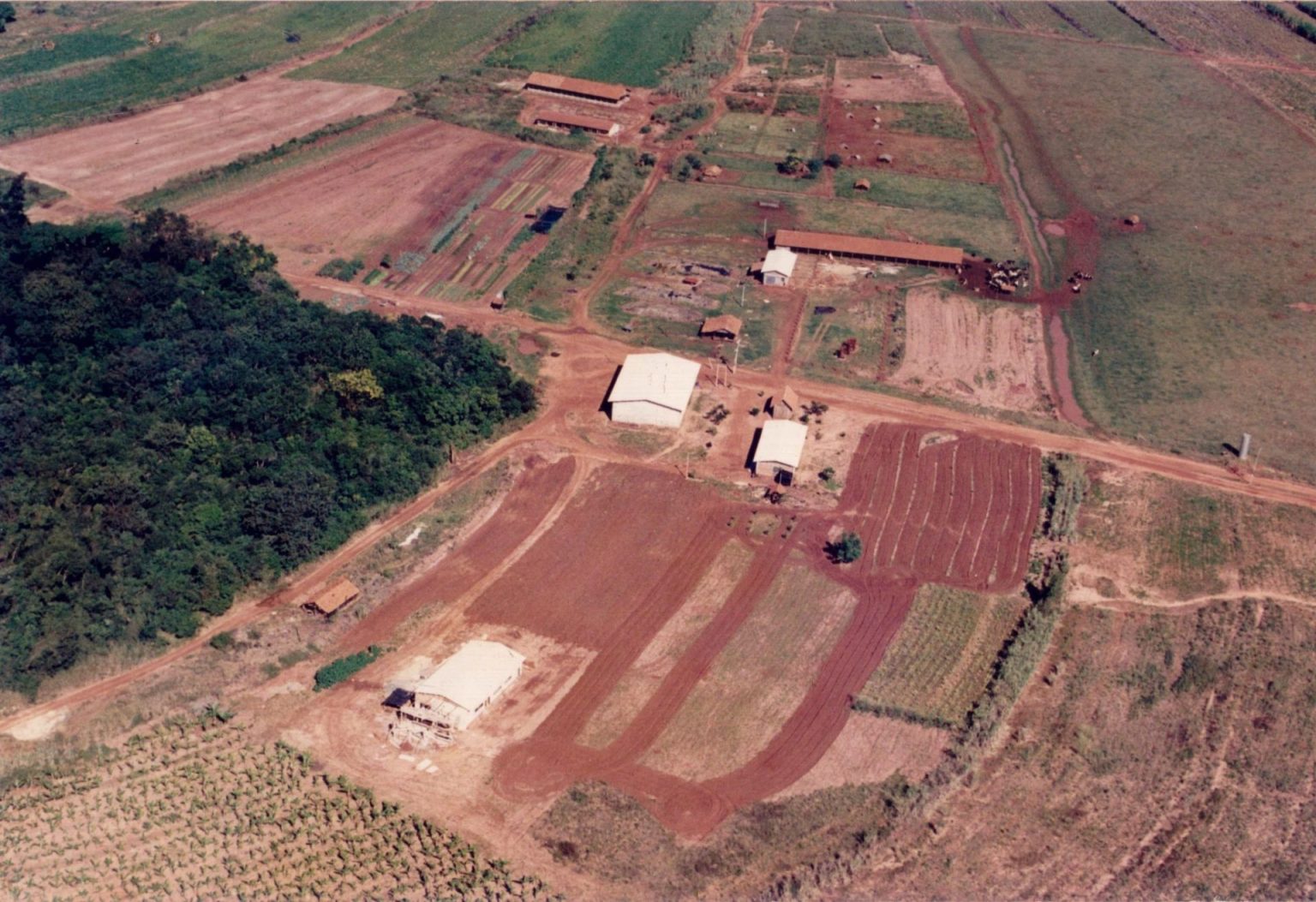 Imagem aérea do assentamento nos anos 90 mostra uma grande área de solo compactado e sem cobertura vegetal, com três estruturas construídas e vestígios de cultivos. À direita, resta uma área de mata nativa