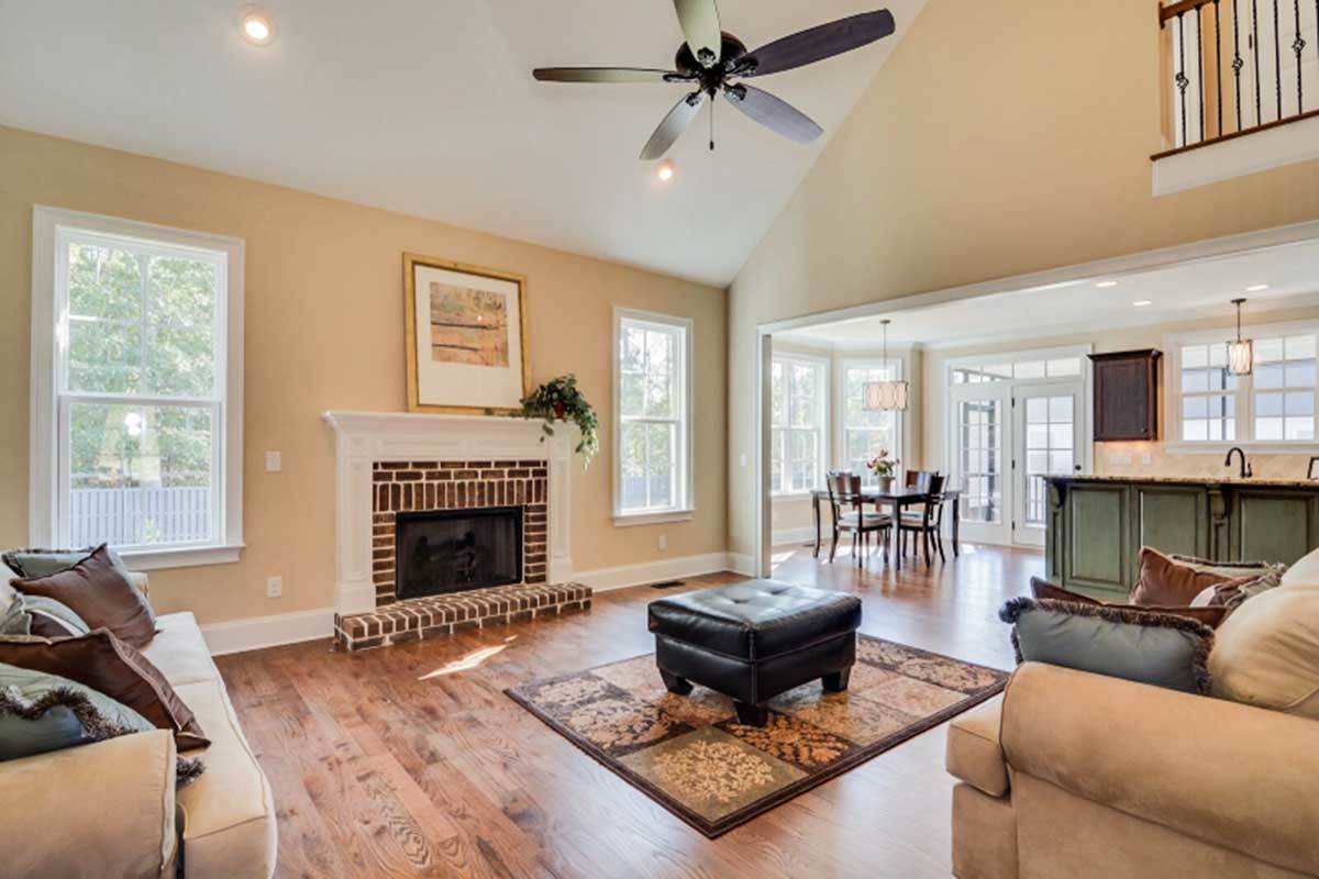 The living room has a vaulted ceiling, beige seats, a tufted ottoman, and a brick fireplace flanked by framed windows.