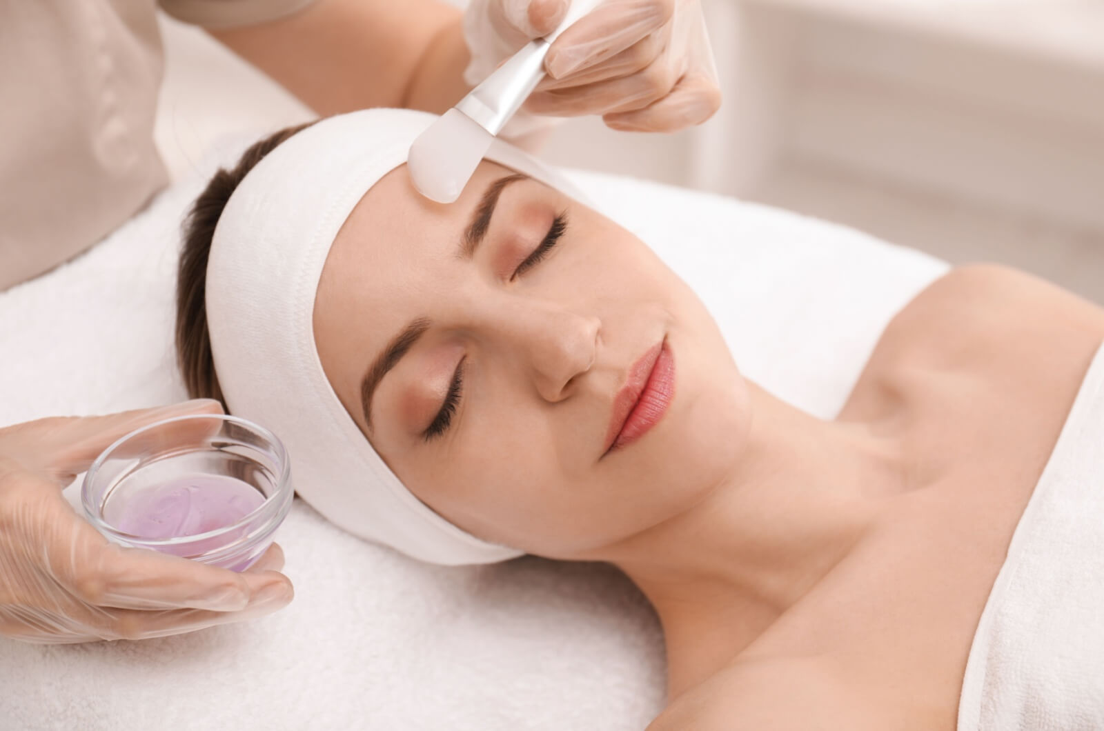 A medical esthetician applying a chemical peel agent with a brush on an individuals face while they are laying on a table with their eyes closed.