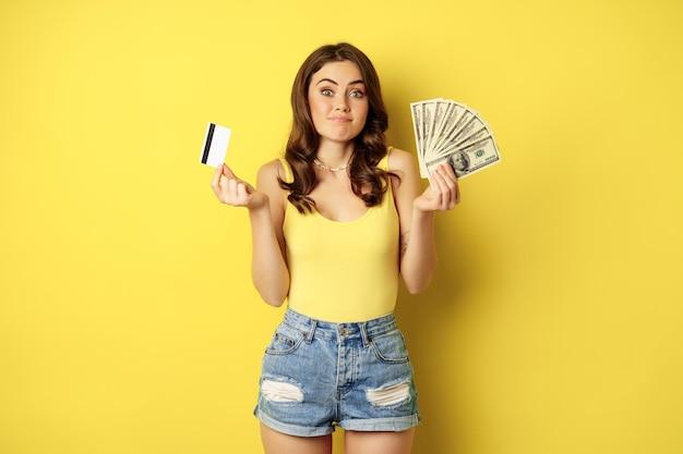 Young pretty woman in summer outfit holding credit card and money cash in hands standing against yel...