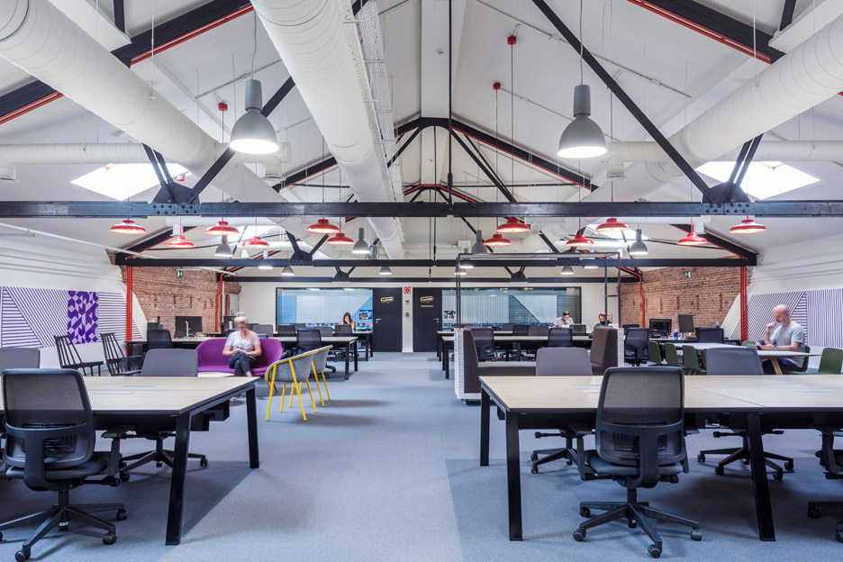 A spacious office with high ceilings and exposed beams showcases industrial adaptive reuse, featuring several desks and office chairs.