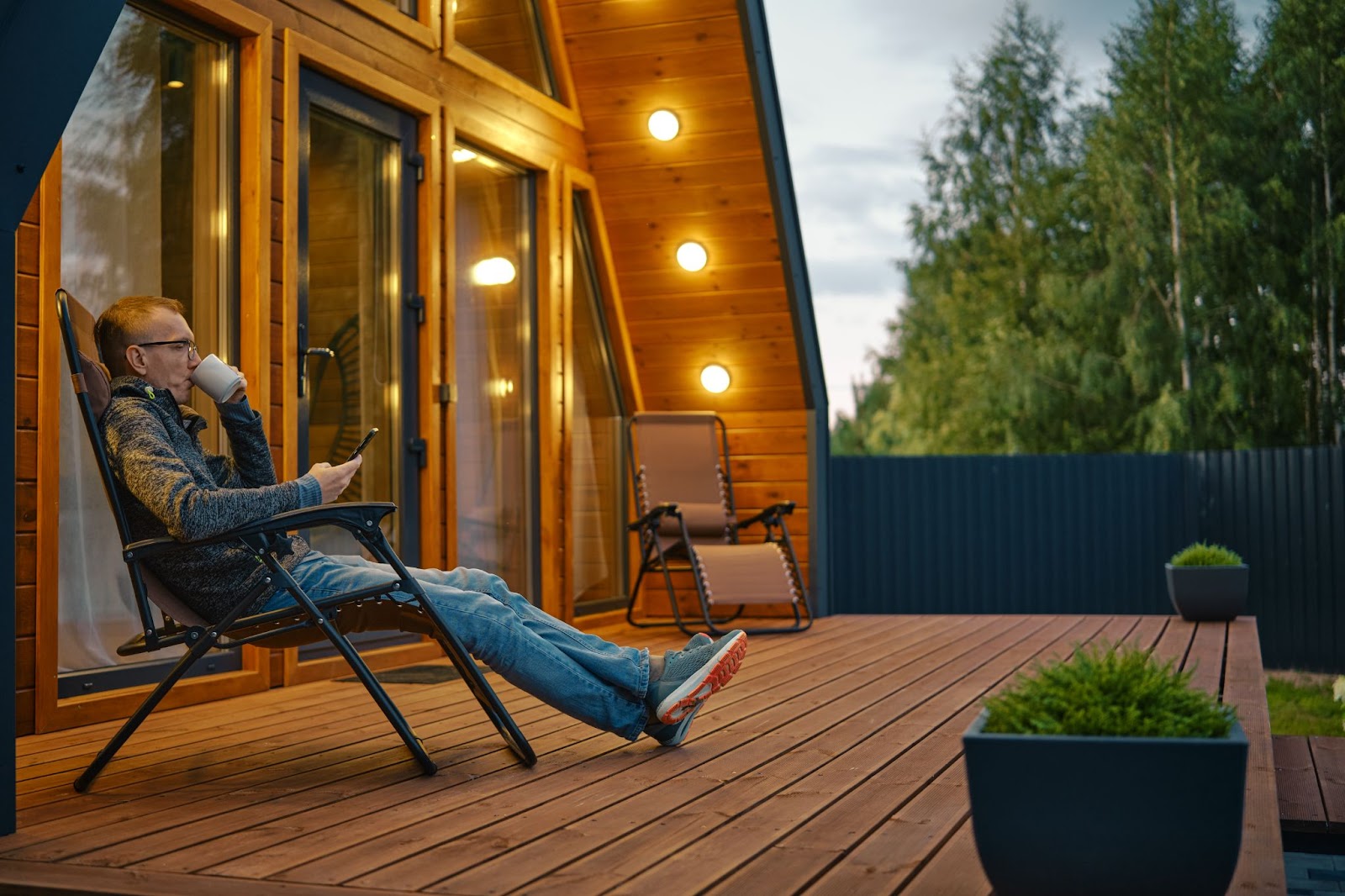 A man sitting on his custom wooden deck outside of his house, sipping on tea and looking at his smartphone.