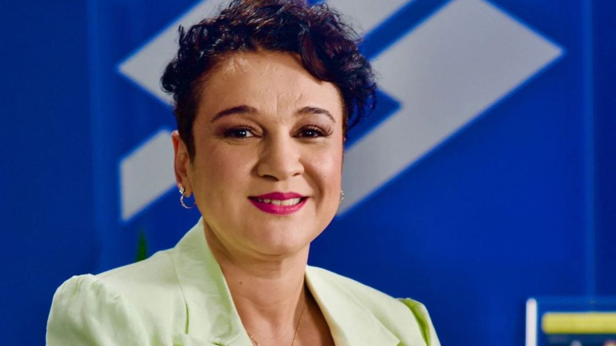 Tarciana Medeiros, presidente do Banco do Brasil, está parada em frente a um fundo azul, sorrindo para a camêra. As suas costas o símbolo do  Banco do Brasil. 