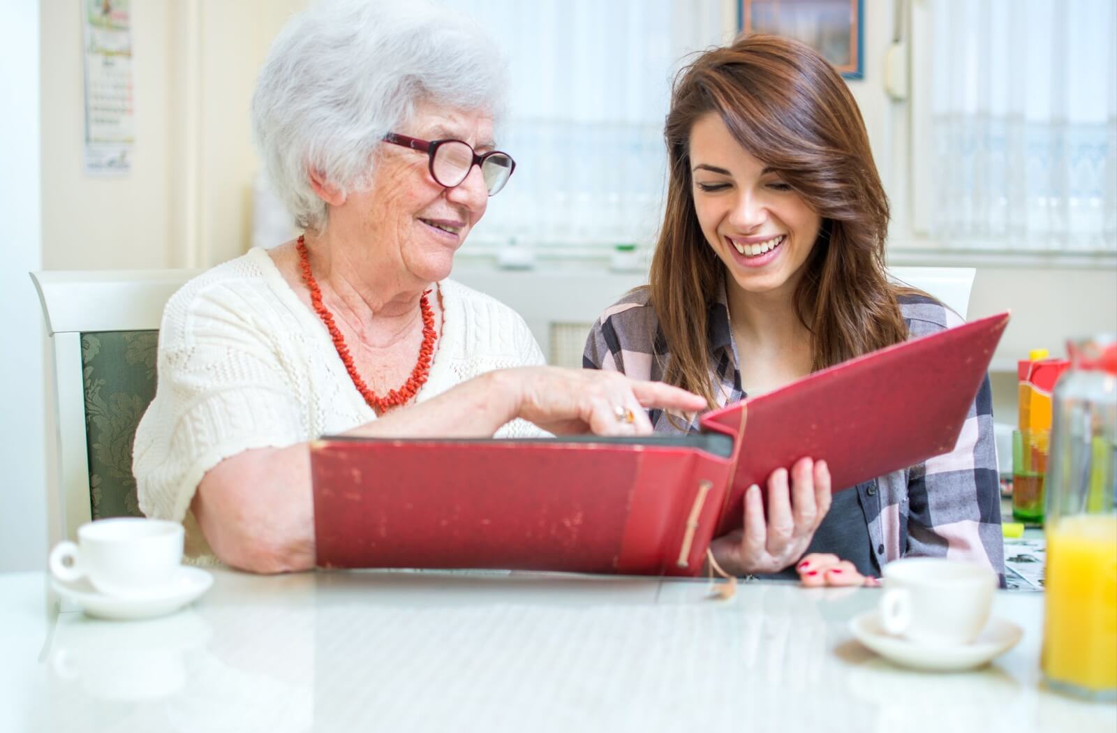 Senior and younger adult looking through a photo album, laughing together at a dining table with coffee and juice nearby