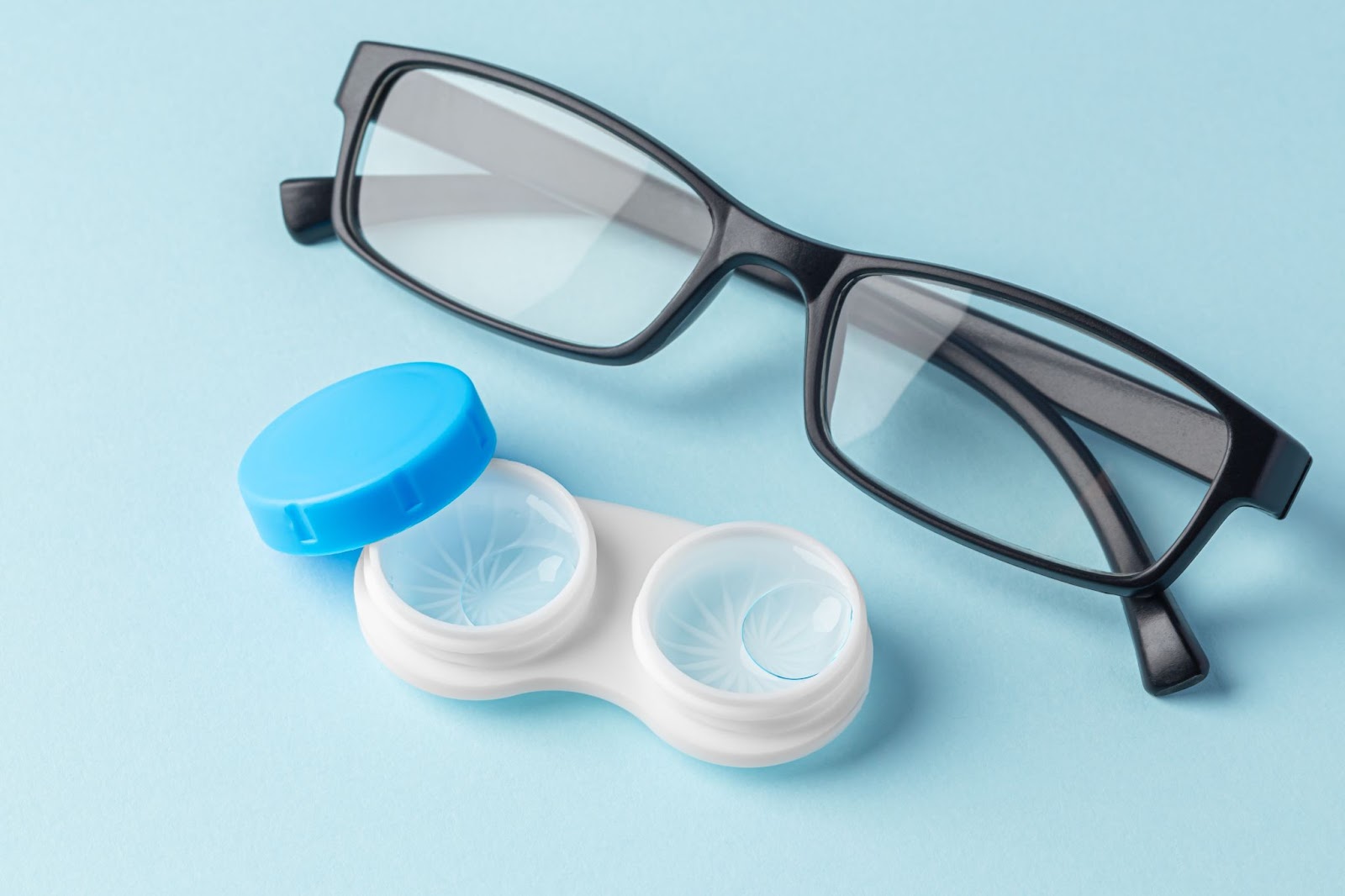 A pair of glasses and a contact lens case, opened to show the lenses inside, sit beside each other on a table.