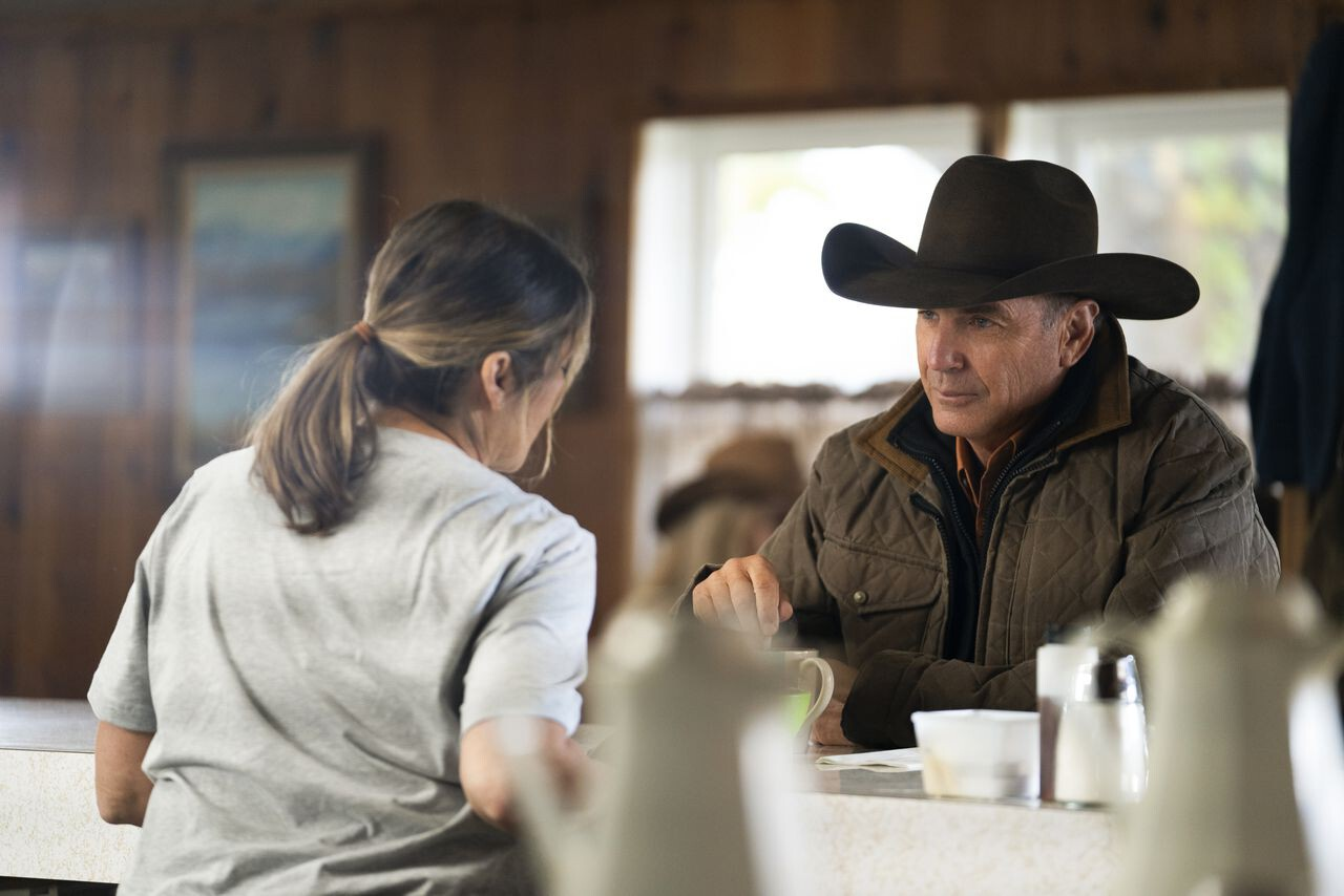 Hombre con sombrero de vaquero sentado en la barra de un restaurante rústico, conversando con una mujer de espaldas.