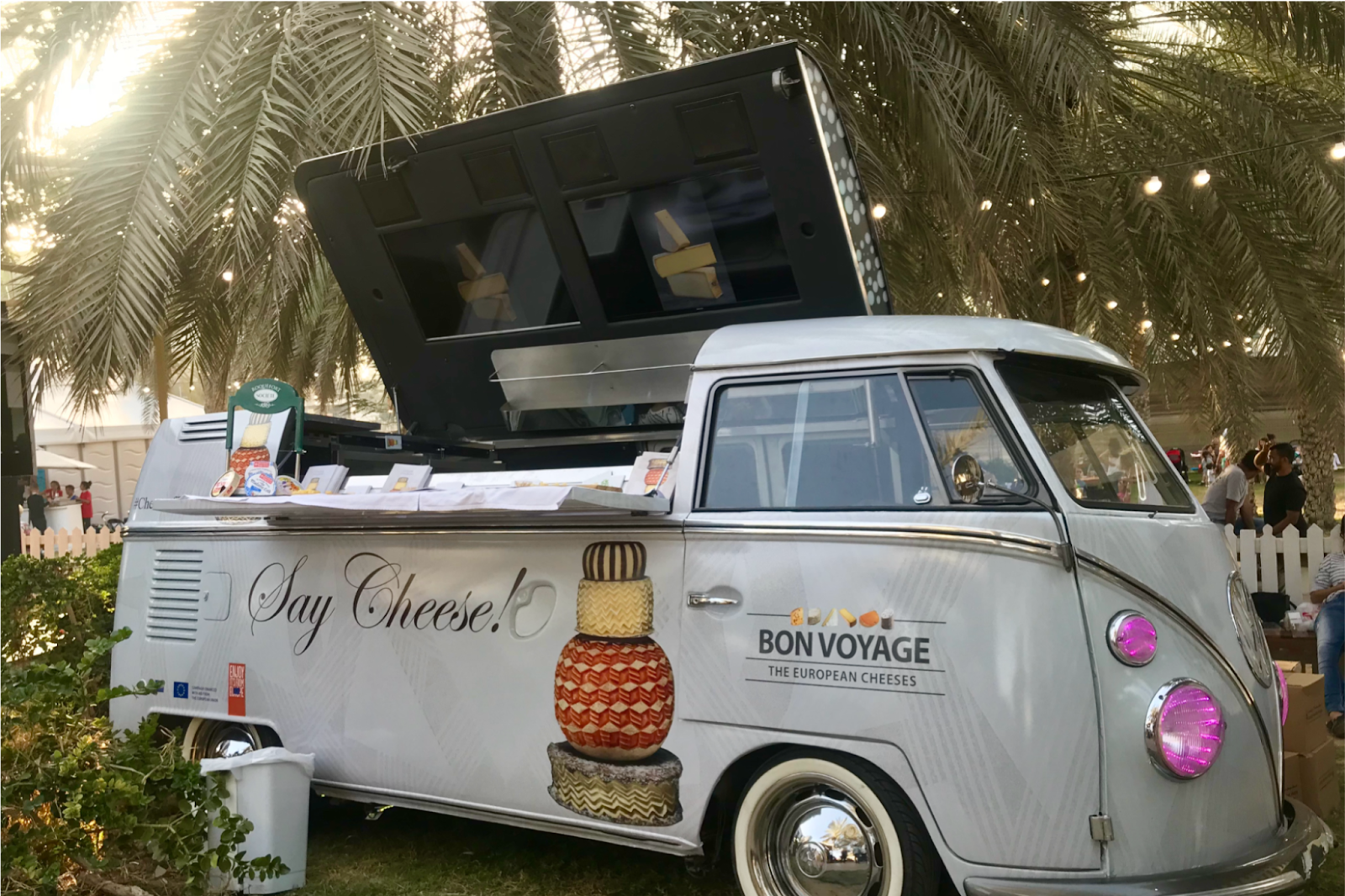Outdoor event venue with a vintage food truck serving guests under palm trees and festoon lighting.