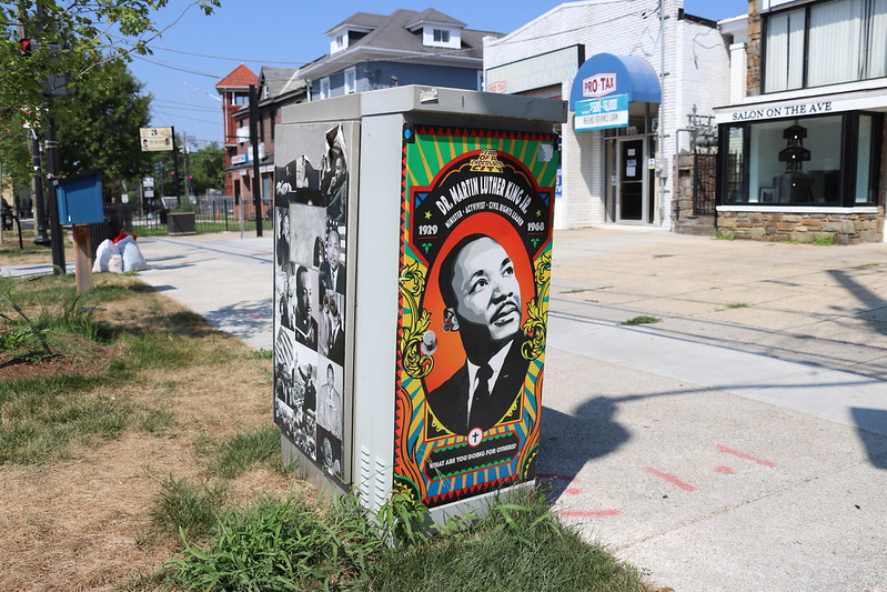 Red, black, gold, and green poster with Martin Luther King's face is on a poster plastered on a utility box in a city block.