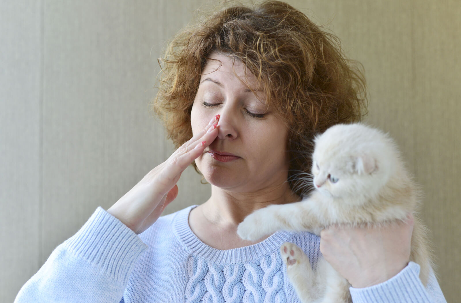 An adult holding a small white kitten and rubbing their eyes in discomfort and irritation.