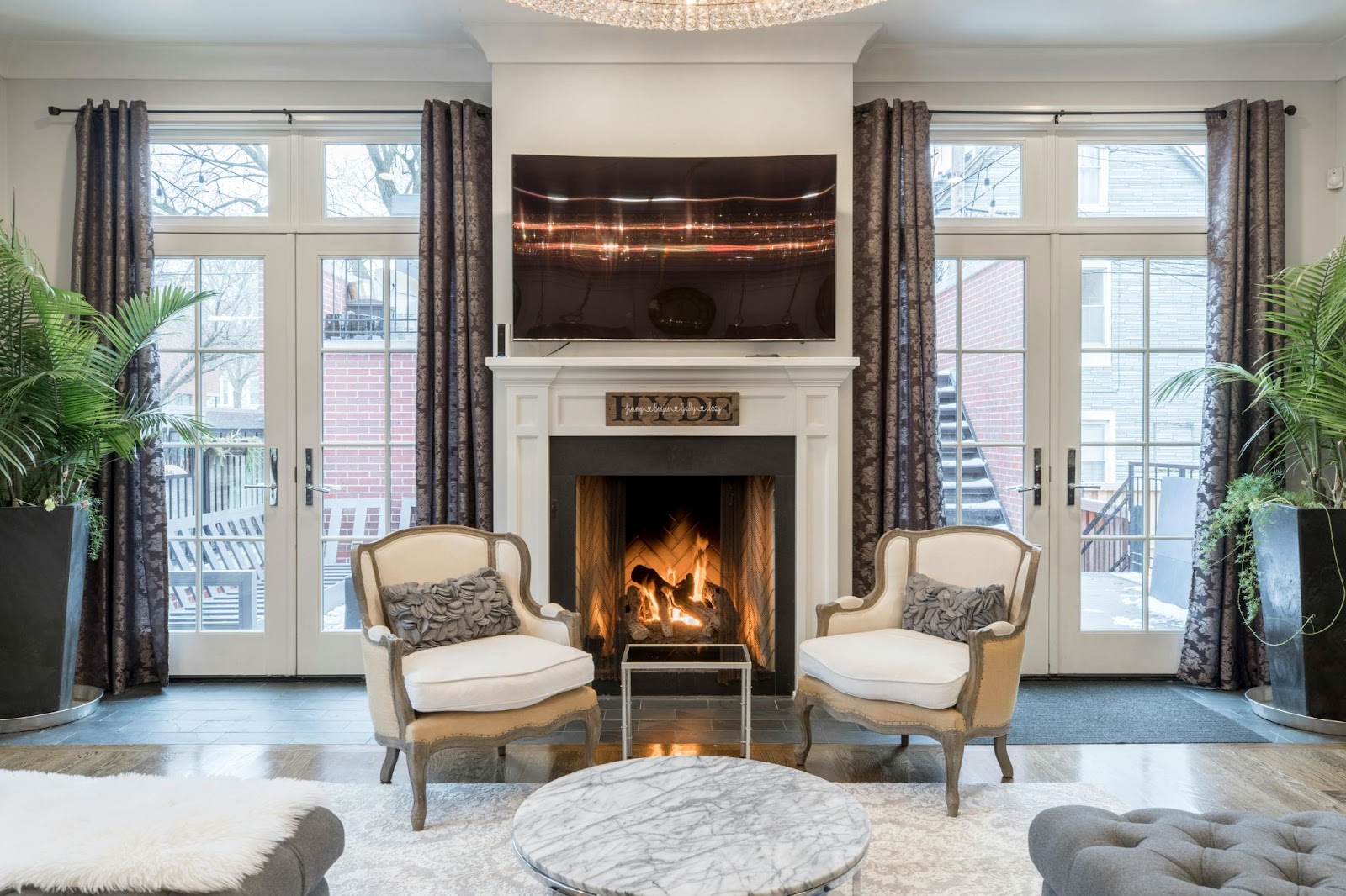 This image depicts a pair of elegant French doors in a modern home. The living area has a fireplace, two lounge chairs and a television on the mantle.