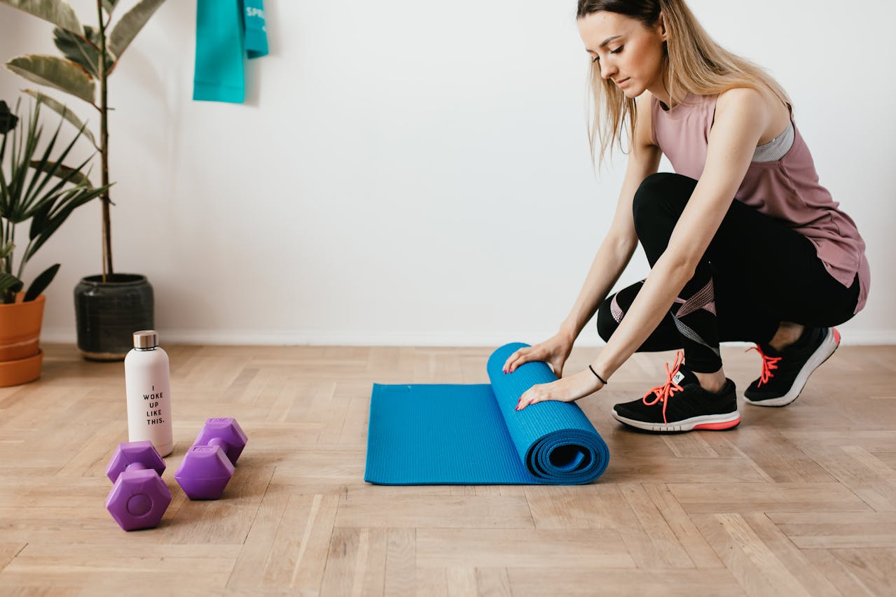 Femeie tânără pregădindu-se să facă sport cu o saltea și gantere la ea acasă. 