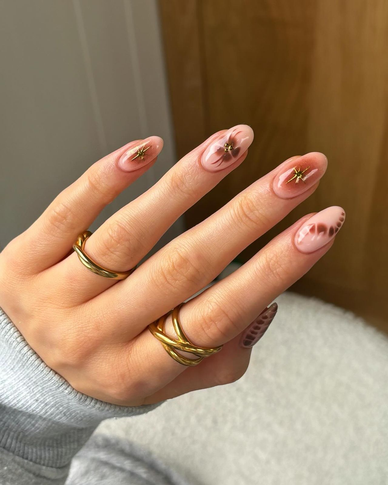 Hand with pink manicured nails adorned with star and dot patterns, wearing gold rings, against a soft background.

