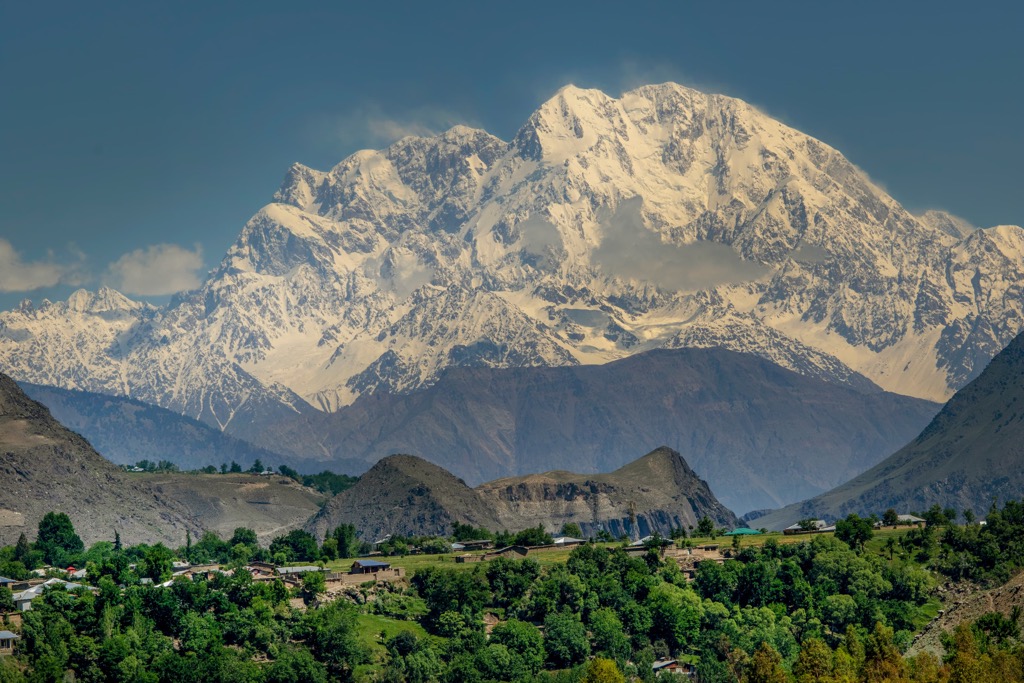 Scenic view of a majestic snow-capped mountain range with a lush green valley and small village in the foreground, under a clear blue sky, where hindu kush is grown.