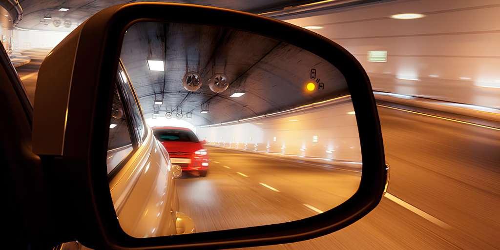  A car's side mirror with the blind spot monitor warning light illuminated, alerting the driver to a red car in the adjacent lane while driving through a tunnel.
