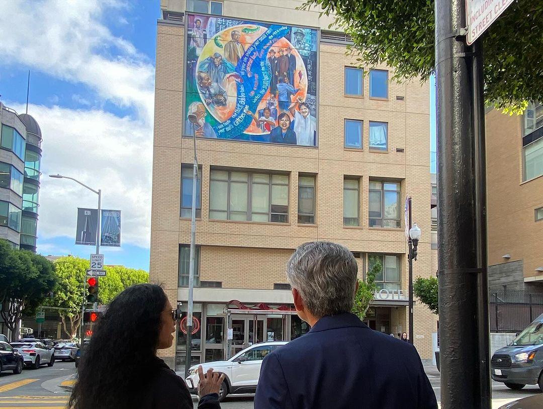 A person and person standing on a sidewalk in front of a mural on a building

Description automatically generated