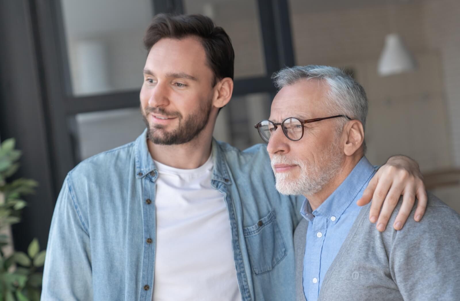 A relaxed caregiver embraces their parent with one arm while looking out the window.