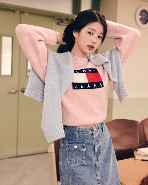 This contain IVE Wonyoung wearing a pink sweater and denim skirt standing in front of a wooden table