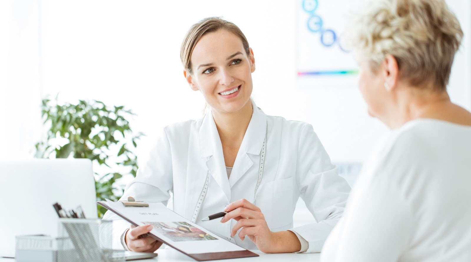 A smiling female registered dietitian in a white coat discusses a health plan with an older female patient in a bright office.