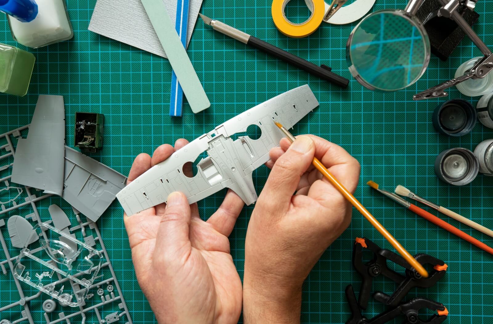 Overhead view of hands painting model airplane wings on a grid desk with many tools and parts.