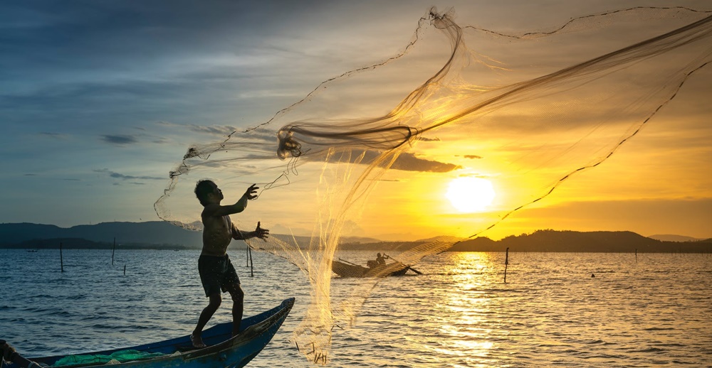  A fisherman stands on the edge of a small wooden boat, casting a large fishing net into the water at sunset, with golden light reflecting off the waves and silhouetted hills in the background.









