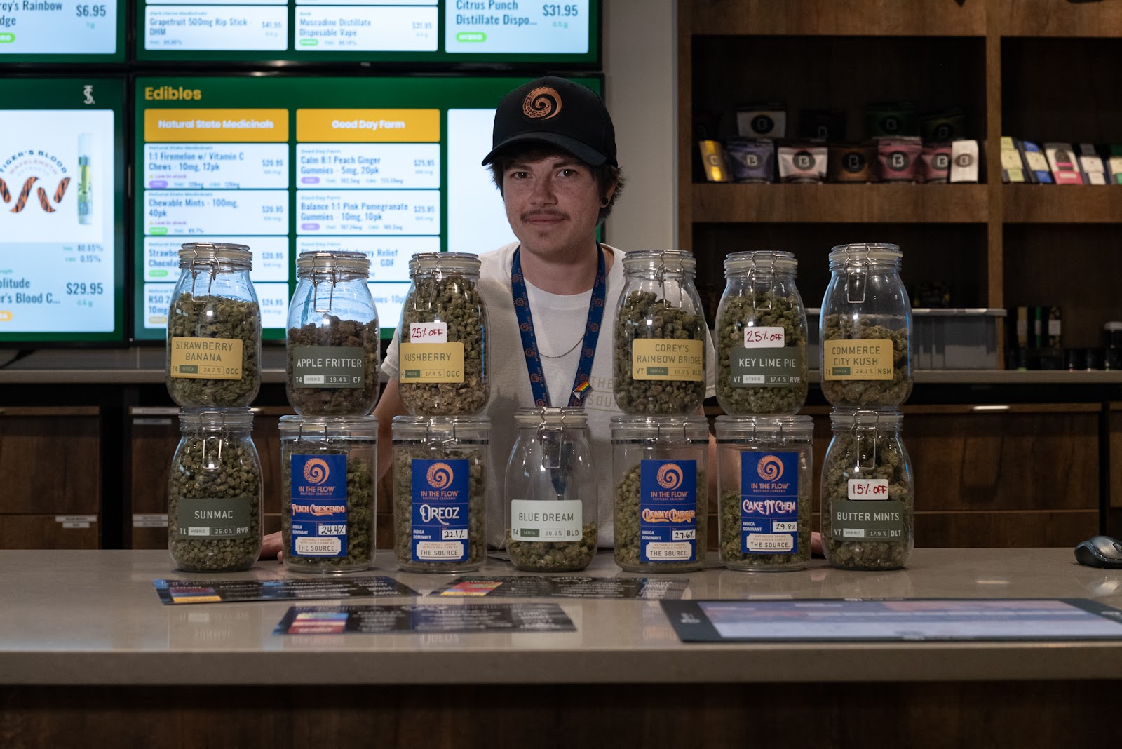 A young budtender wearing an In The Flow boutique cannabis hat stands between a couple of rows of jars filled with cannabis flower. Six jars on top, seven jars on the bottom, the jars display a wide variety of cannabis strains with varying THC percentages.