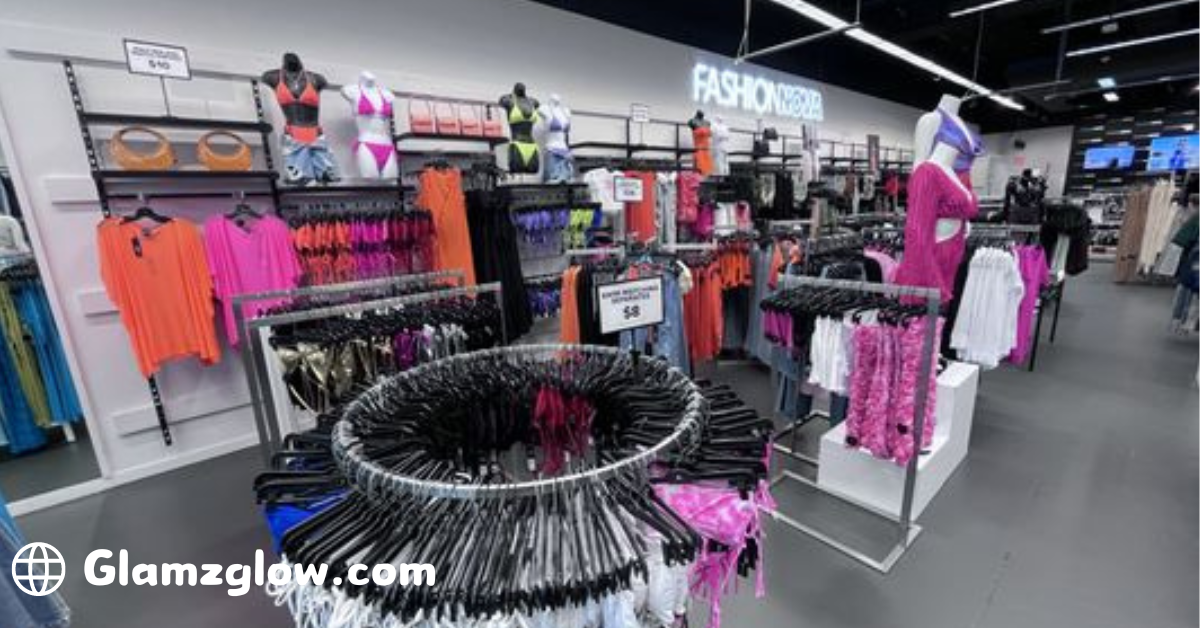 The image shows the interior of a Fashion Nova store, filled with racks of colorful clothing. Various outfits, including swimsuits, dresses, and tops in vibrant colors like pink, orange, and purple, are displayed. Mannequins wearing stylish outfits are placed throughout the store. In the background, a large illuminated sign that reads "FASHION NOVA" is visible. At the bottom left corner, the website URL "Glamzglow.com" is displayed with a globe icon next to it.