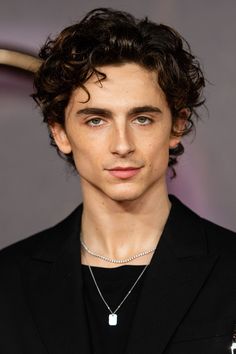 This contain Timothée Chalamet   with curly hair wearing a black suit and diamond necklace on his neck