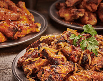 A plate of crispy, charred chicken wings topped with caramelized onions, served with fresh parsley garnish alongside other wing varieties.