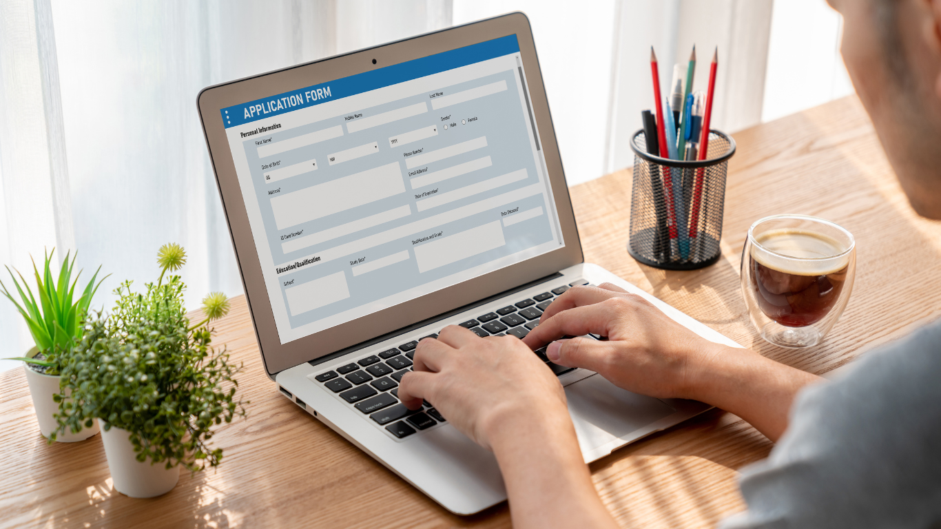 Person completing online application form on laptop computer at modern home office workspace with plants