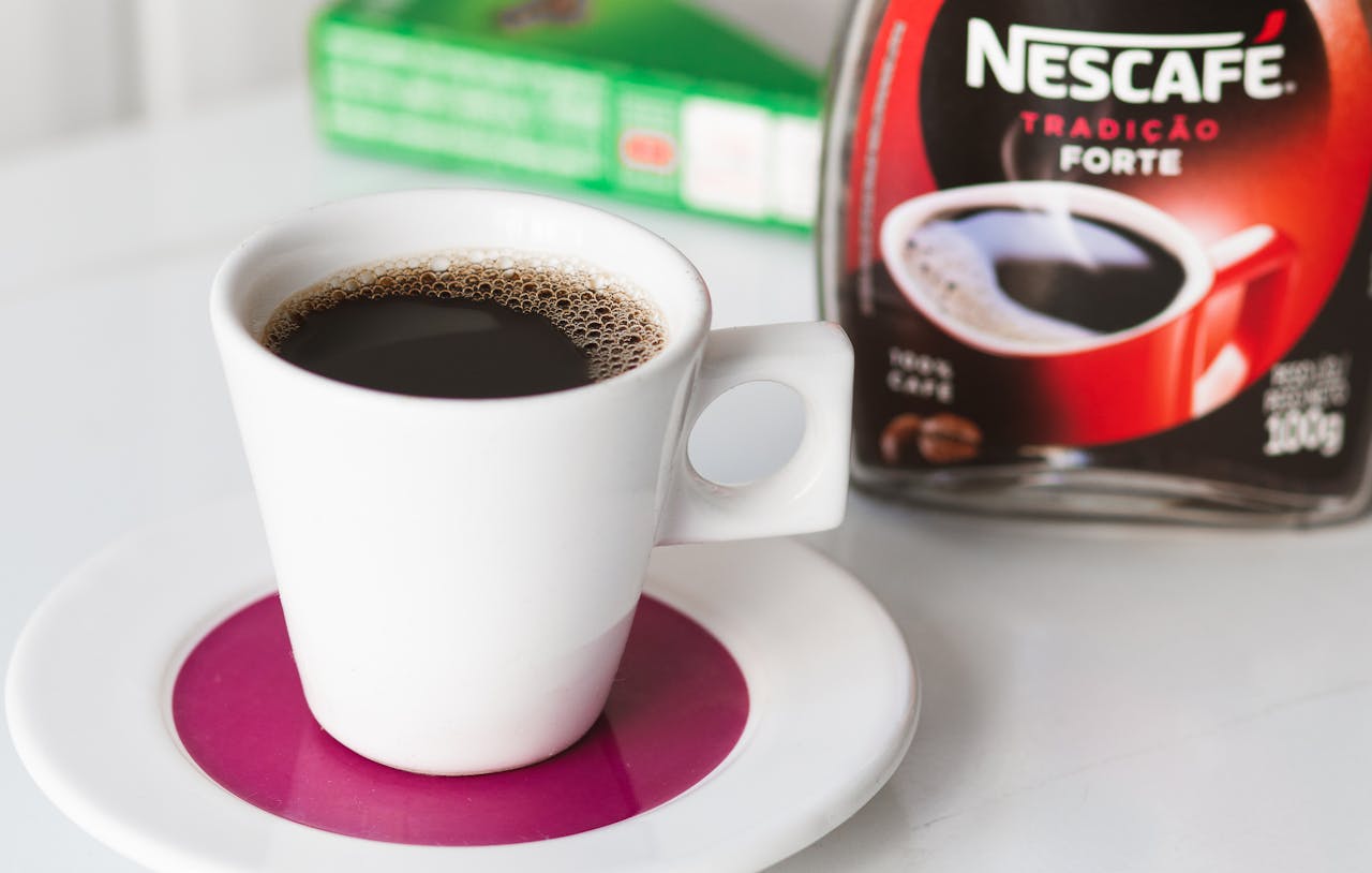 The image shows a white cup filled with black coffee, placed on a saucer with a purple center. In the background, a jar of Nescafé Tradição Forte instant coffee is partially visible. The focus is on the freshly brewed coffee, with tiny bubbles on the surface, creating a simple yet appealing coffee setup.