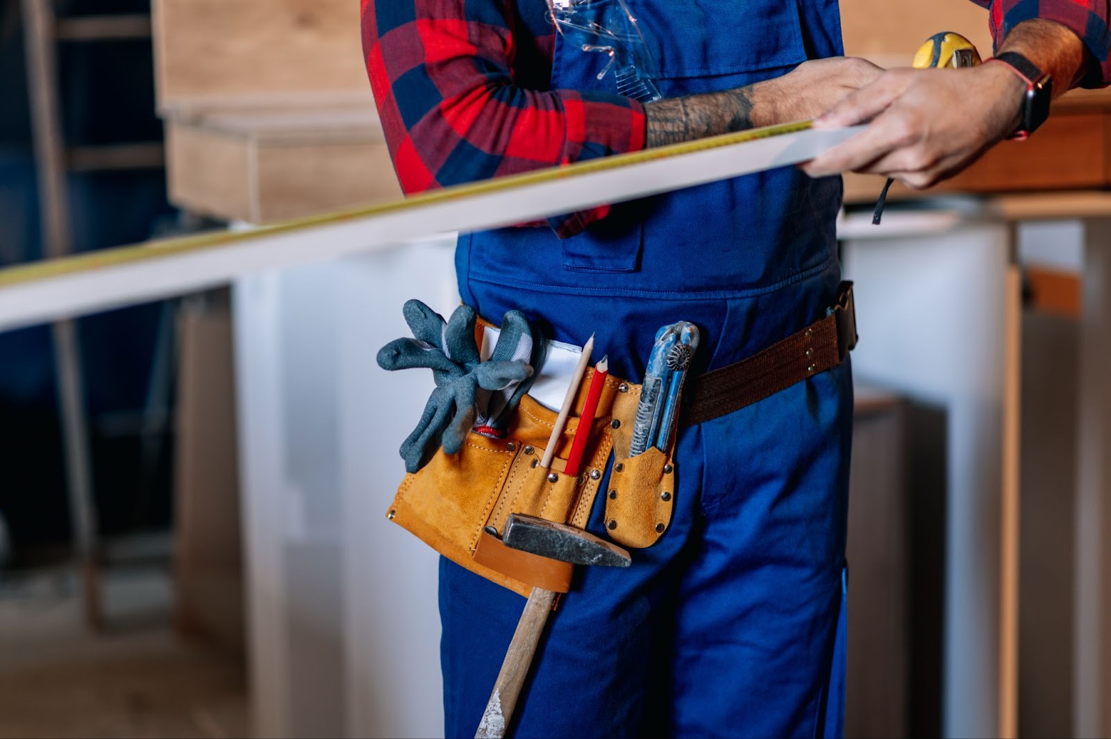 A close-up of a young male handyman measuring wood with a tape measure, wearing a tool belt with various tools around his waist.