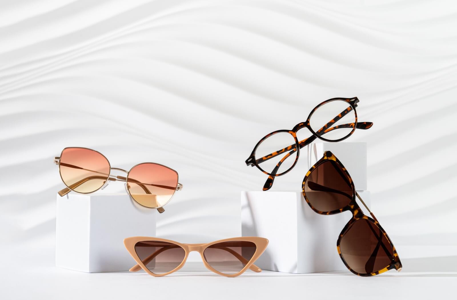 Four stylish glasses on white blocks with minimalist wave background featuring tortoiseshell frames cat-eye sunglasses and gradient lens eyewear.