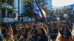 This contains a large group of people walking down a street holding flags and rainbow colored skirts in the sun