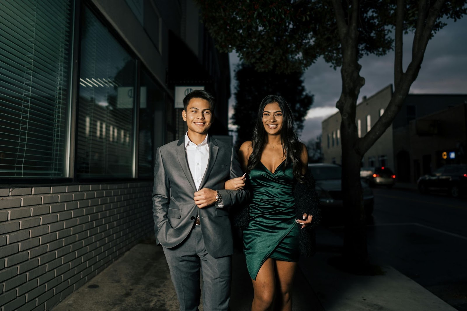 a couple is walking down the street. he is wearing a suit and she a pretty dress. They are both smiling.