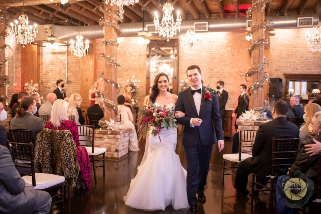 In the historic, industrial chic Grand Hall of Butler's Courtyard smiling newlyweds joyfully make their exit after their wedding ceremony. The couple's happiness is palpable as they walk through the venue, surrounded by the distinctive architectural features. This is one of many perfect wedding ceremony locations in League City Texas, near Houston Texas. Guests can stay cool and dry!