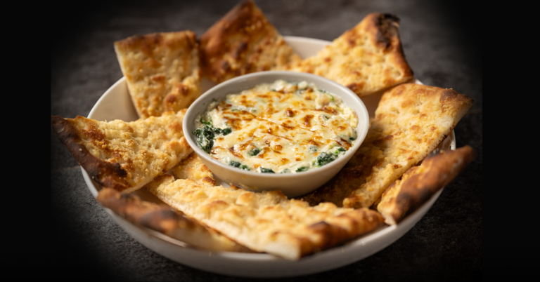 A warm spinach and artichoke dip topped with melted cheese, served in a bowl with crispy, charred flatbread pieces on a dark table.