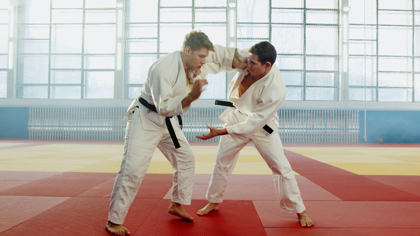 Two martial arts practitioners in white uniforms train together in a well-lit dojo.