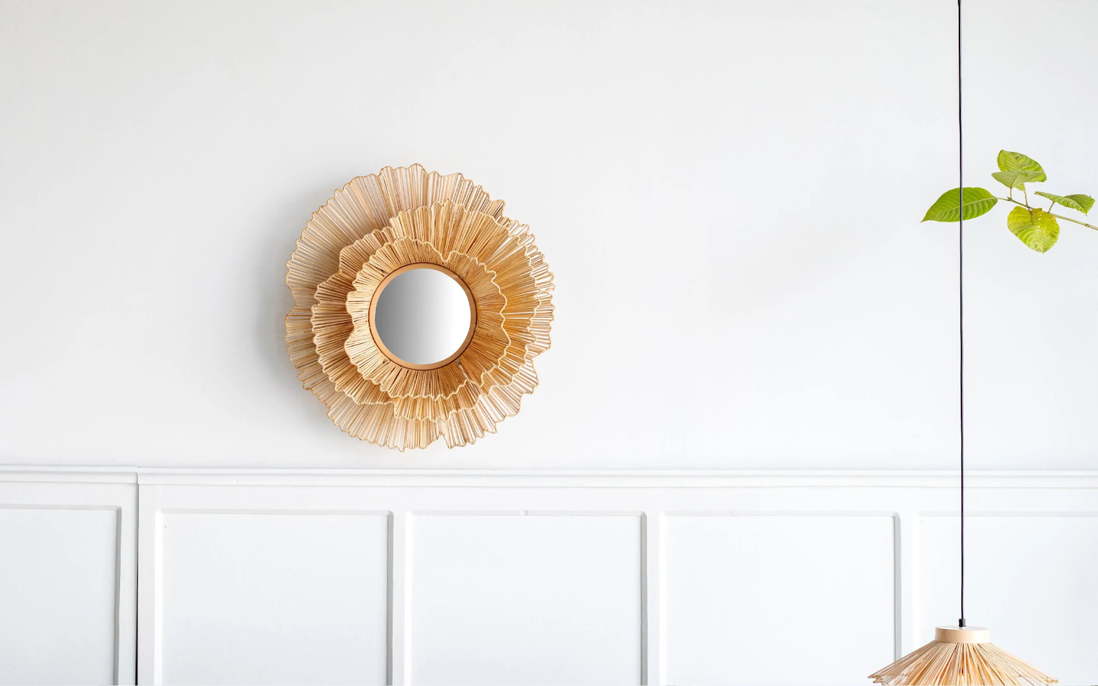 Decorative wall mirror with a layered, flower-like rattan frame mounted on a white wall, with a pendant lamp and a green plant branch hanging on the right side.
