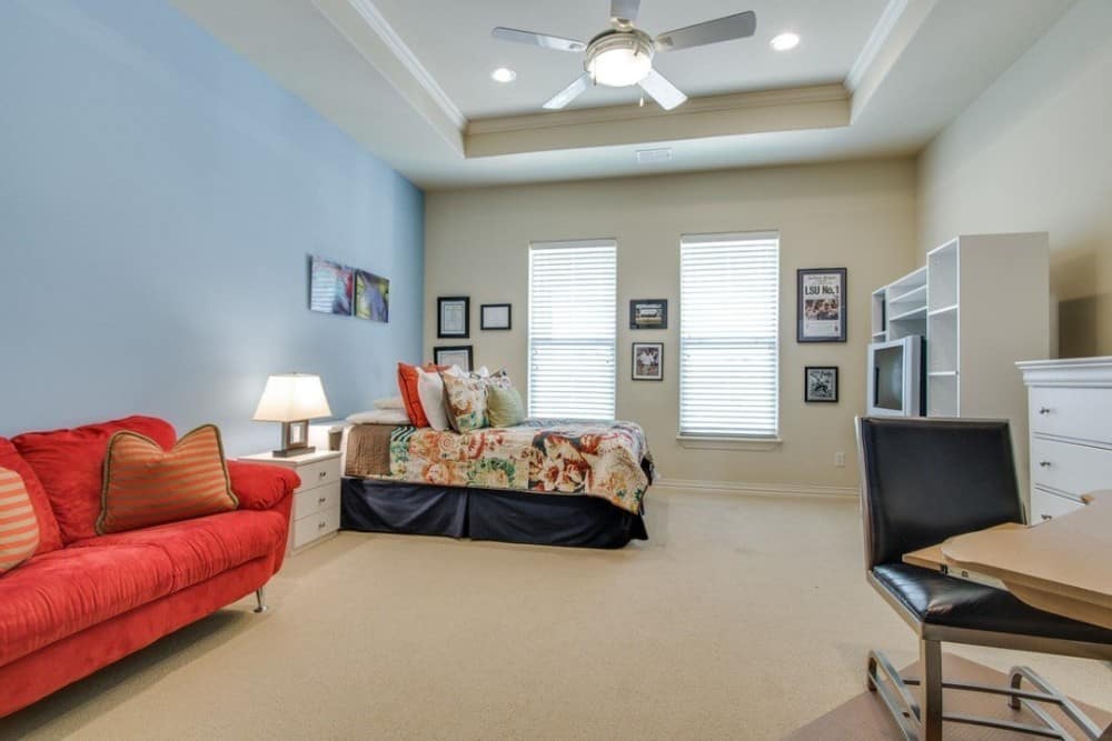 The primary bedroom has a tray ceiling, a skirted bed, and a red sofa over the carpet flooring.