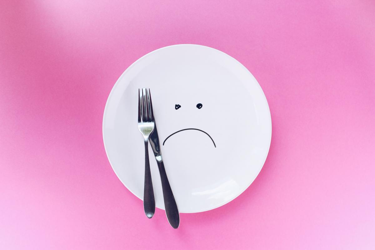 A photo featuring a white plate with a fork and spoon on one side, with a sad face drawn in the center against a pink background.