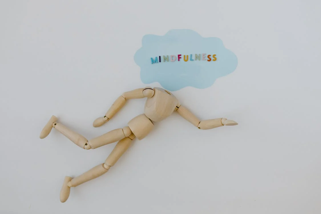 A wooden art dummy without a head. The dummy is in a moving position. Where the head would be is a blue cloud shape made from paper, with multicoloured letters spelling out "mindfulness".