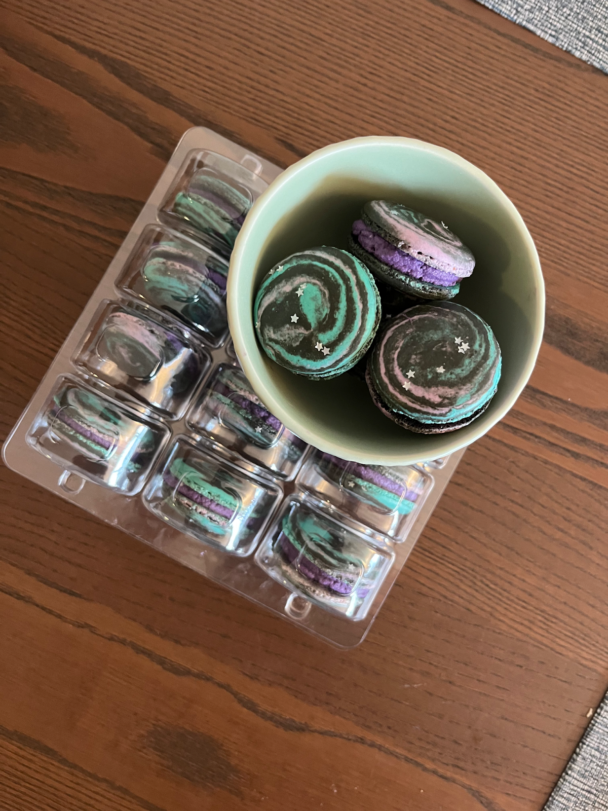 A bowl of galaxy print macarons, dusted with stars, sit on a plastic case filled with more.