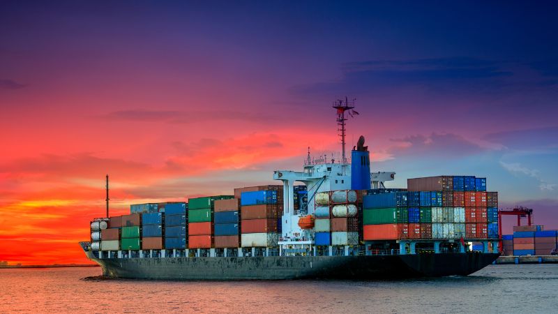 A cargo ship with colorful containers sails at sunset, against an orange and purple sky.