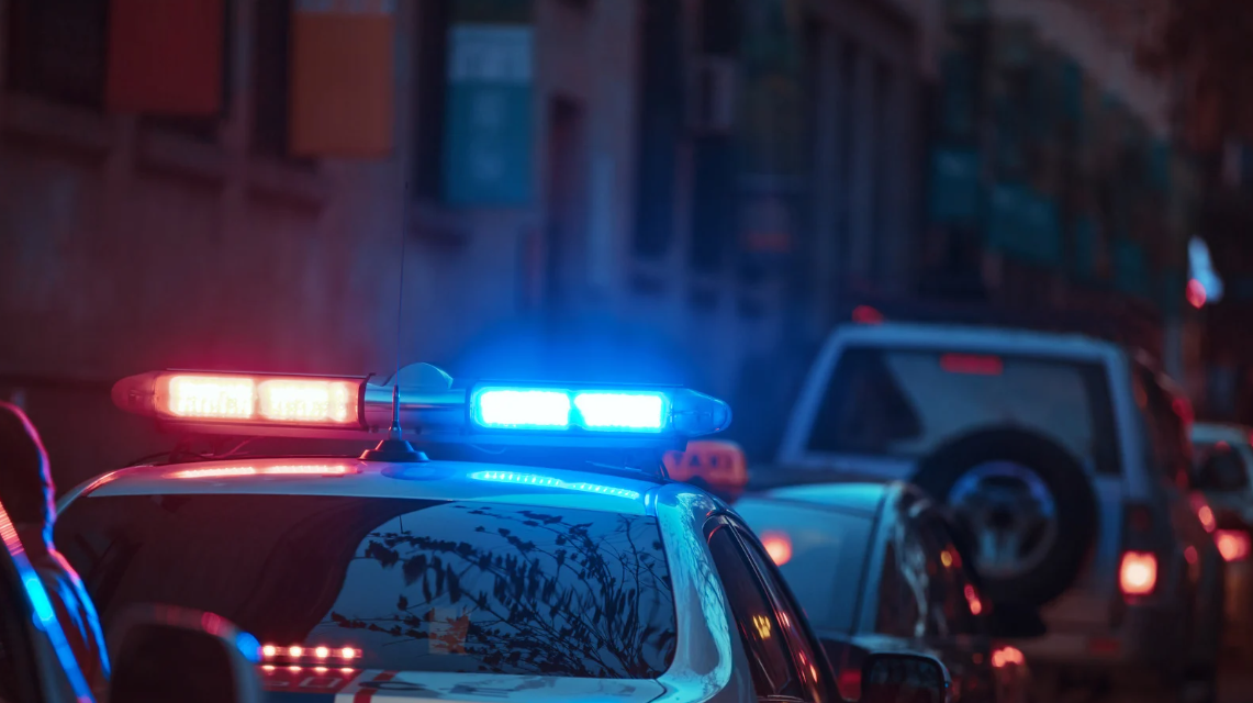 (Close-up of a police car with red and blue sirens flashing on a city street at dusk, indicating an active law enforcement presence or response.)