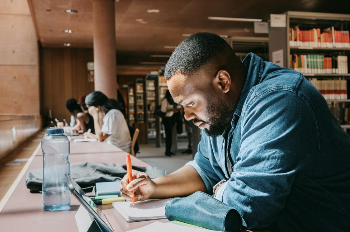 A Black man in a blue shirt writes on a notebook in a library - Double Major Tips