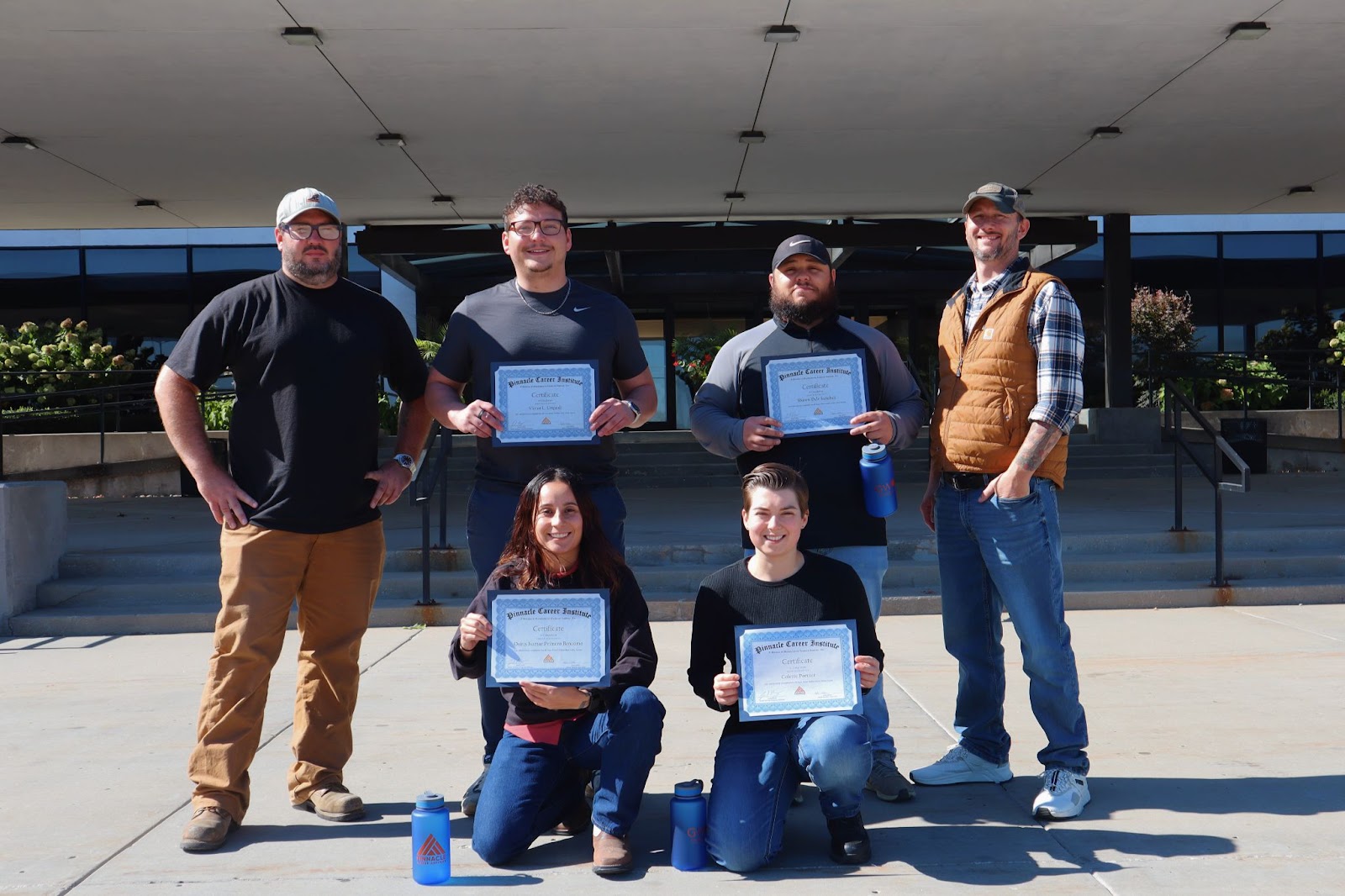 Six individuals, four men and two women, pose outdoors in front of a building, holding certificates of completion for the in-person portion of their Wind Turbine Technician program at Pinnacle Career Institute. Three men stand in the back row, while two women kneel in the front. All participants are smiling, celebrating their achievement.